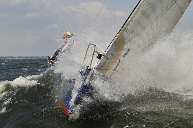 Photograph of a J/133 heavy weather sailing, it's bow is smashing through the crest of a large wave. Overcoming wave resistance